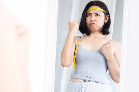 Asian woman motivate herself to lose weight standing in front of a mirror with measuring tape, girl with inspiration for weight loss goalの写真素材