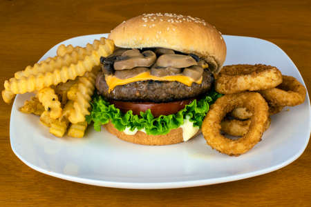 hamburger top with melted cheese and mushrooms with a side of fries and onion ringsの写真素材