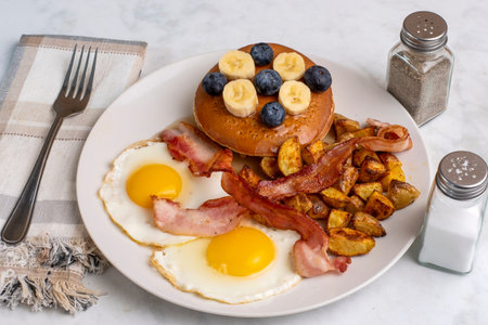 fried eggs with pancakes topped with bananas and blueberries served with home fries and bacon.