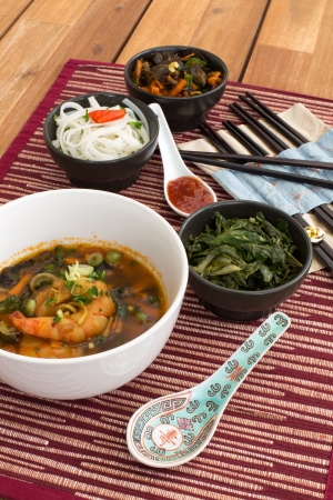 Japanese soup with shrimps in white ceramic bowl composed with ceramic spoon with spicy red sauce, japanese chopsticks and black bowls with rice noodles, kale (green cabbage) and fried vegetables. Composition on a old styled wooden table.の写真素材