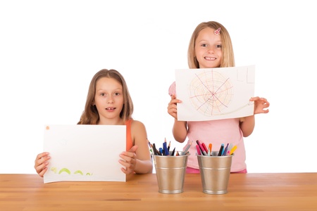 Two sisters with blond hair smiling and showing their super drawings at a wooden table with metallic pencils holderの写真素材
