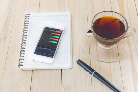 view of stock market application on touchscreen smartphone with coffee, book and pen on wooden...