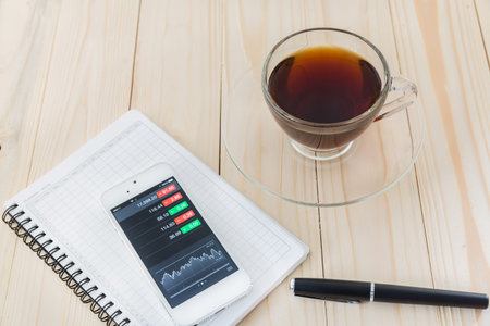 view of stock market application on touchscreen smartphone with cup of coffee and a book