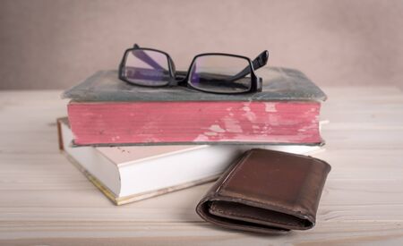 brown wallet and stack of old books, reading glasses on a wooden table-selective focus on walletの写真素材