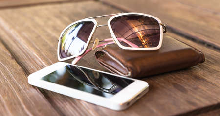 a set of smartphone and tinted sunglasses atop a brown leather wallet on wooden background - vintage effect styleの写真素材