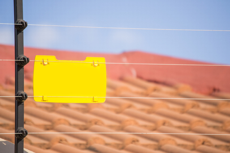 Elecric fence istalled on a wall with a blank yellow sign and a blurred roof of a house in the backgroundの写真素材