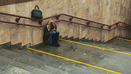 Young girl on the stairs in the subway depressed sitting alone obscures the face with his hands and...