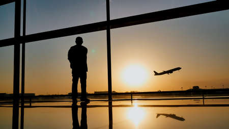 Silhouette of a tourist guy watching the take-off of the plane standing at the airport window at sunset in the evening. Travel concept, people in the airport.の写真素材