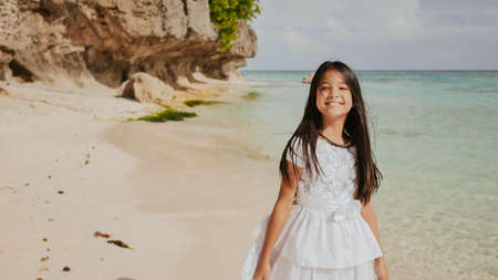 A charming and happy philippine teenage girl in a white summer dress is running along a tropical beach near the rocks. She is happily spinning. Childhood. Recreation.の写真素材
