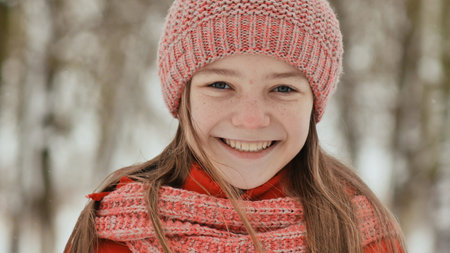 Portrait of a young schoolgirl with freckles in the woods in winter. He warms his hands in mittens and applies them to his face and lips. Shows movement hand in hand.の写真素材