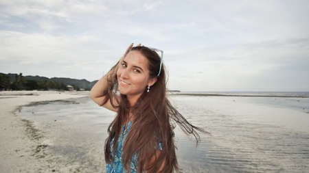 A beautiful girl with long hair in a long blue dress strolls along the beach. Girl posing on camera on exotic coast. Shooting in motion.の写真素材