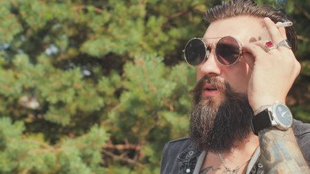 A bearded man with big tattoos smokes a cigarette in the street.