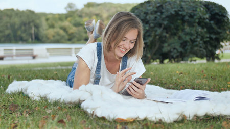 A young girl lies on a white plaid in the park and dials a message on the phone.