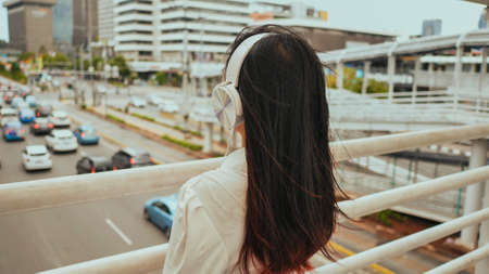 Asian girl stands on a bridge over road traffic and in white headphones listens to music.の写真素材