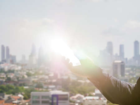 A man holds a phone in his hand that shines a ray of sunshine against the background of the city of Jakarta. Indonesia.の写真素材
