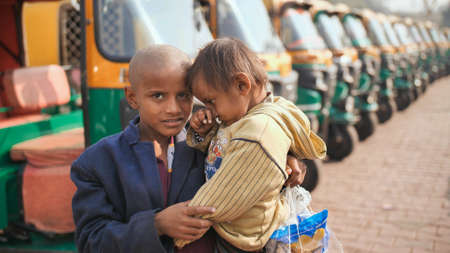 Agra, India - December 12, 2018: A little beggar boy holds a baby in his arms against the background of a rickshaw.のeditorial素材