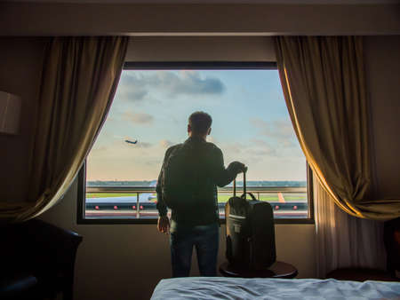A young guy looks at the plane taking off from the window of his hotel room.の写真素材