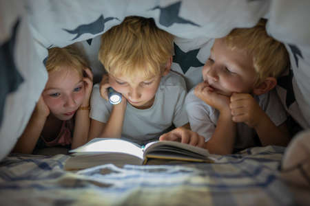 Three young children are reading a book with a flashlight under the covers at night.の写真素材