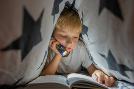 A little boy reads a book with a flashlight under the covers at night.の写真素材