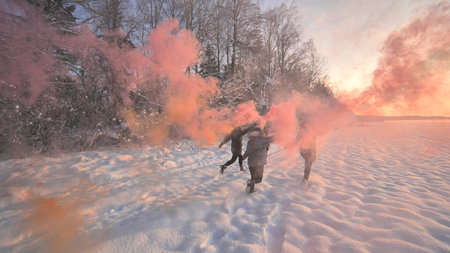 Girls friends run across the winter field with colored smoke.