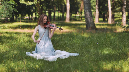 A young girl plays the violin on the sitting grass in the park.の写真素材