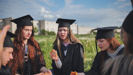 Graduates in black suits eating pizza in a city meadow.の写真素材