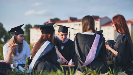 Graduates in black suits eating pizza in a city meadow.の写真素材