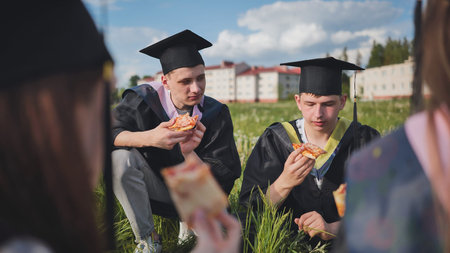 Graduates in black suits eating pizza in a city meadow.の写真素材