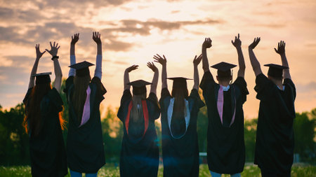 Silhouettes of graduates in black robes waving their arms against the evening sunset.の写真素材