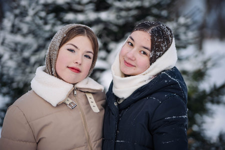 Two young schoolgirls wearing winter clothes are smiling in a snowy forest during winterの写真素材