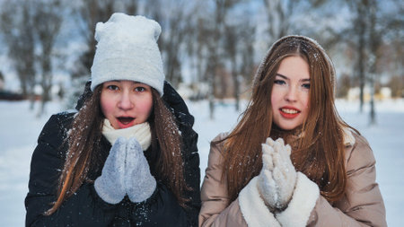 Two young schoolgirls in winter attire, smiling and warming their hands in a snowy parkの写真素材