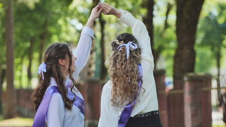 Two students in graduation sashes joyfully dance together in the schoolyard to celebrate their last...