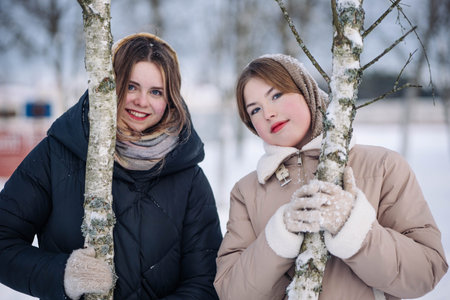 Two smiling schoolgirls are posing with birch trees in a snowy winter park, enjoying their leisure timeの写真素材