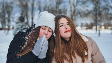 Two schoolgirls standing in a snowy park, warming hands during winter snowfall, bundled up in warm clothing. Happy and cheerful, showcasing friendship in the coldの写真素材