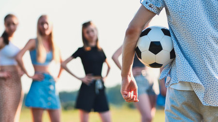 Young man is holding a soccer ball with his friends standing behind him out of focus on a sunny day