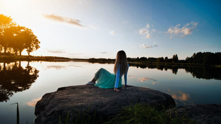 Blonde woman relaxing on lakeside rock, basking in golden sunset glow, serene water mirroring cloudy sky backdropの写真素材
