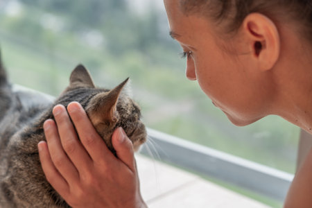 Gentle woman petting fluffy cat, sharing peaceful connection near sunlit windowの写真素材