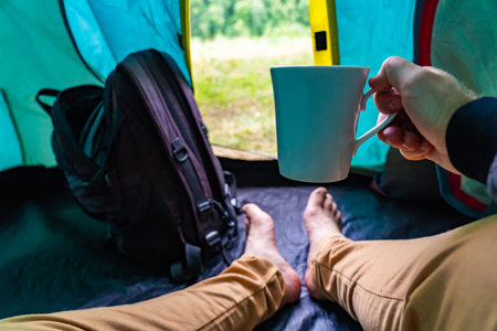 Hiker sipping warm drink inside camping tent, resting after mountain trail adventure
