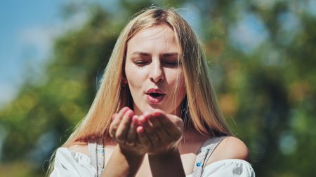 Blonde woman softly blowing kiss, lush green background, sunlight warming her faceの写真素材