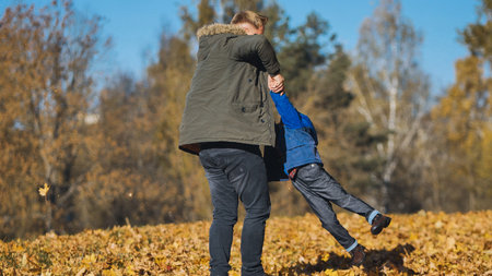 Father and son playing, laughing together among golden autumn leaves scattered across sunny park ground, creating warm family memoryの写真素材