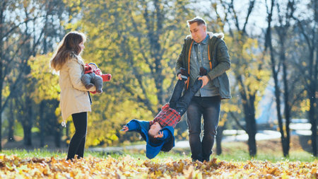 Happy family enjoying a sunny autumn day in a park, father holding his son upside down while mother holding baby, surrounded by colorful fallen leavesの写真素材