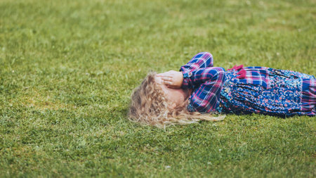 Sad child hiding face, resting on verdant lawn during bright summer afternoonの写真素材