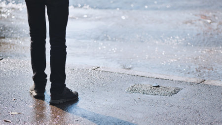 Close up of a mans legs and feet standing near a water leak on an asphalt road, observing the flowing waterの写真素材