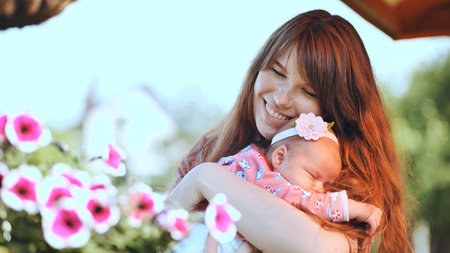 Smiling mother holding her sleeping baby girl, enjoying a peaceful moment in a garden full of flowers, celebrating family dayの写真素材