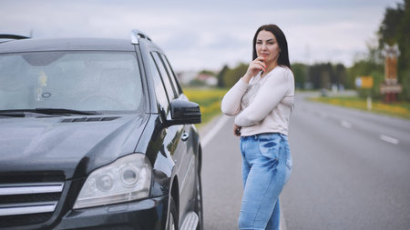 Professional female executive standing beside luxury vehicle near rural highway, contemplating with...