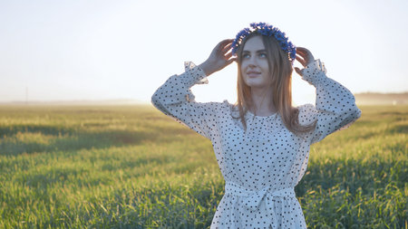 Young woman with flower wreath adjusting her hair while standing in a field at sunset, enjoying the peace and beauty of natureの写真素材