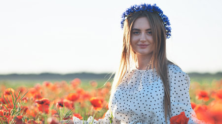 Young woman wearing cornflower wreath, standing gracefully in poppy field, embodying summers natural beauty and floral eleganceの写真素材