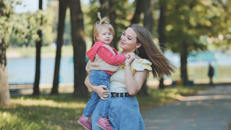 Cheerful mom raising laughing child high during playful outdoor moment amid green park landscape bathed in warm sunlightの写真素材