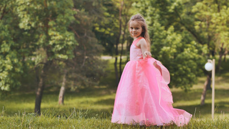 Young girl twirling in pink princess dress, strolling through verdant park during sunny dayの写真素材