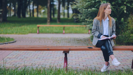 Young woman reading book, relaxing on urban park bench, surrounded by greenery and soft sunlightの写真素材